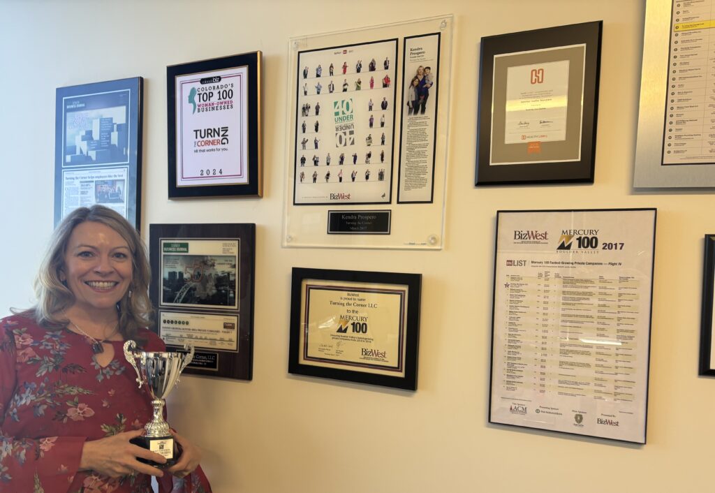 Kendra Prospero, Founder and CEO of Turning the Corner HR, standing in front of the company's office awards wall featuring business awards, community recognition, and organizational achievements.